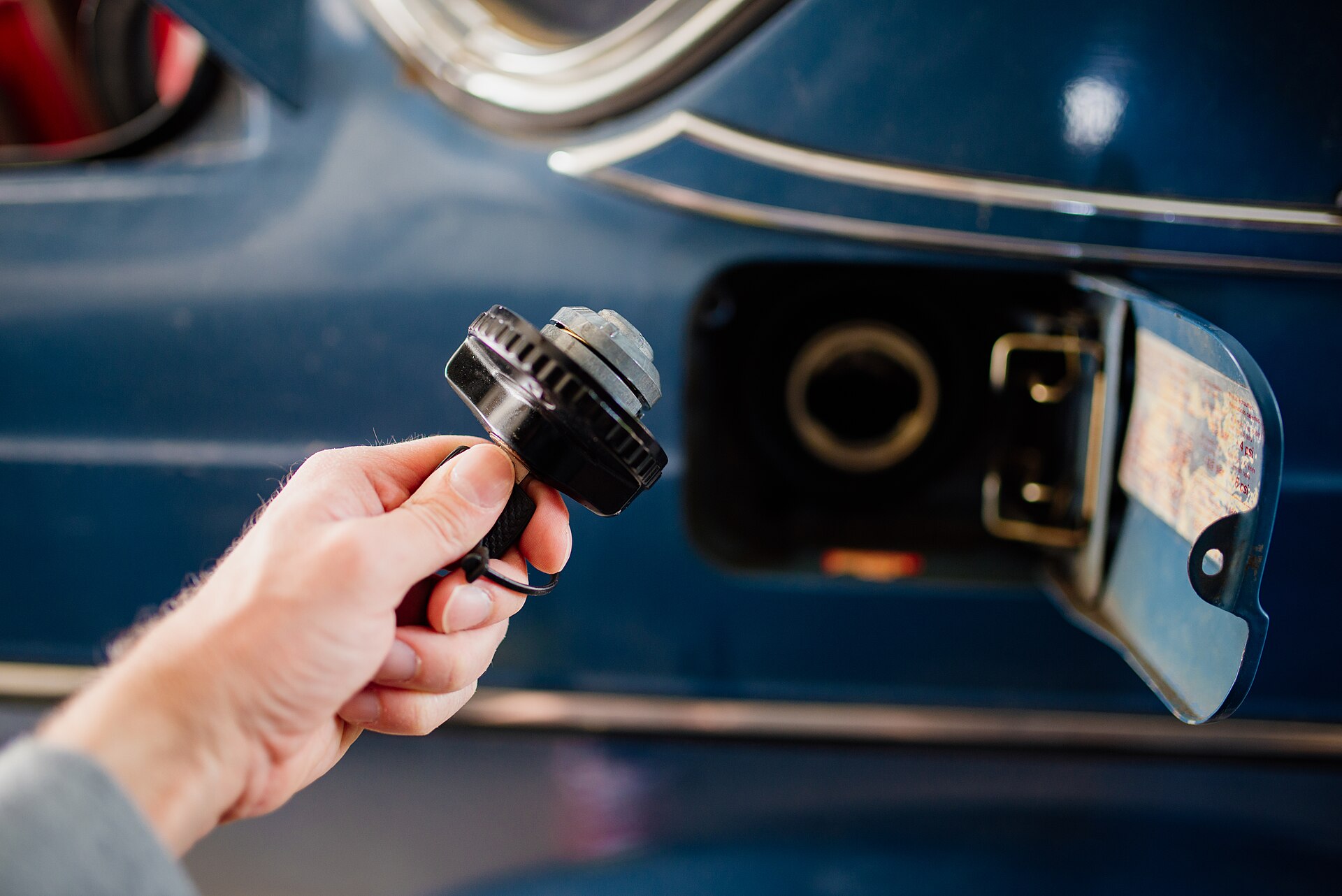 Hand holding a fuel cap beside a car's open fuel filler at a gas station