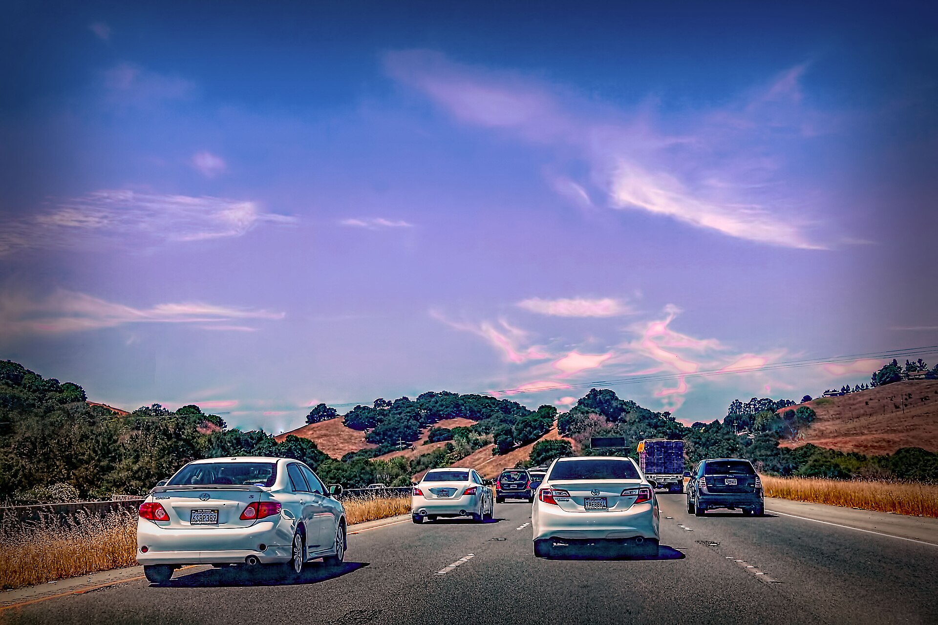 Cars driving along a highway through dry rolling hills under a purple-blue sky